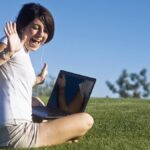 On the left-hand side is a lady with white top and khaki shorts out in the field with her laptop in the background are two green trees and blue sky. There lady is excited to have her laptop working.